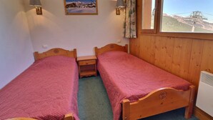 Une chambre avec deux lits en bois, une petite table de chevet, une lampe fixée au mur, une fenêtre donnant sur une montagne et un tableau accroché au mur.