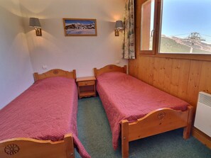Una habitación con dos camas de madera, una mesita de noche pequeña, una lámpara de pared, un ventanal con vista a la montaña y un cuadro en la pared.