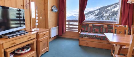 A cozy room with a wooden interior, a large window with a view of snow-covered mountains, a wooden bench with a patterned cushion, and a flat-screen TV on a wooden stand.
