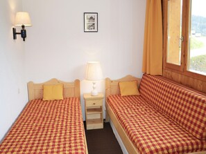 Ein Zimmer mit zwei Betten, jeweils mit kariertem Überwurf, einem Nachttisch mit Lampe, einem gerahmten Bild an der Wand und einem Fenster mit Vorhängen.