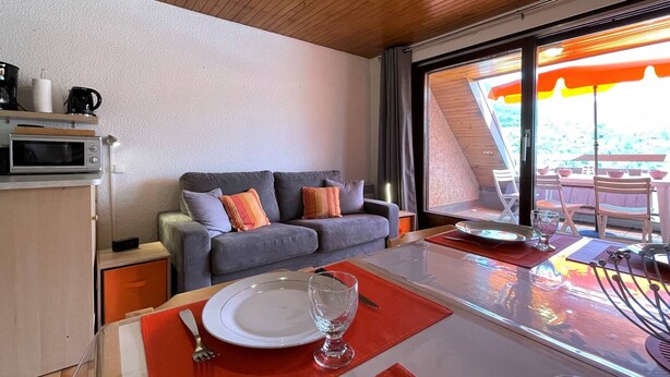 A modern room with a wooden ceiling, a grey sofa with colorful cushions, a dining table with a red placemat, and a view of an outdoor area through a large window.