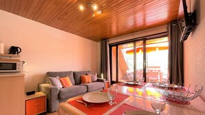 A cozy room with a wooden ceiling, a grey sofa with colorful cushions, a dining table set with red placemats, and a large window with curtains.