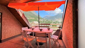 A balcony with a table and chairs, a red umbrella, and a view of a mountainous landscape.