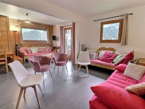 A cozy cabin interior with a wooden floor, a pink sofa, a wooden bed, and a dining area with chairs and a table.
