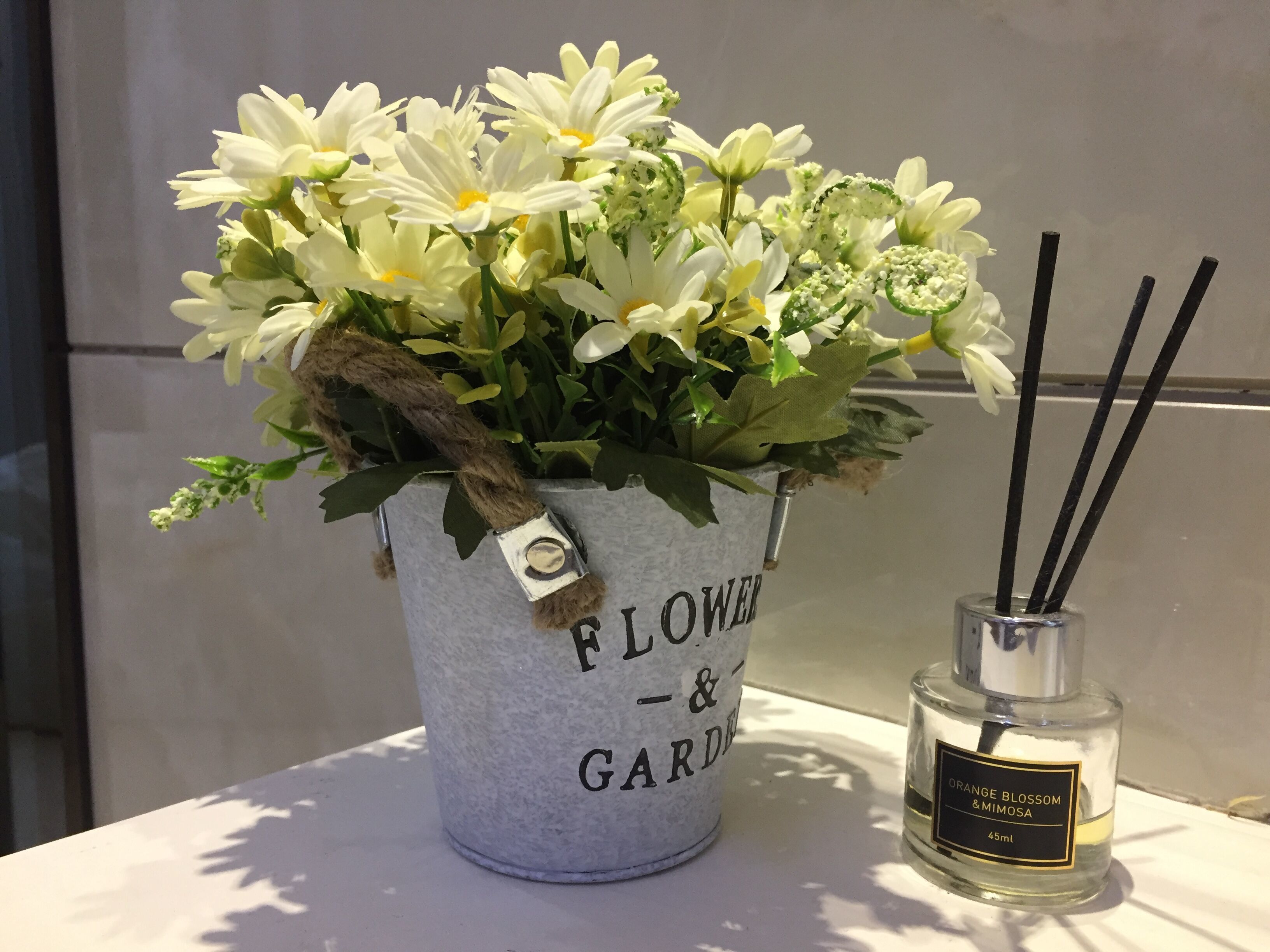 A flower arrangement in a bucket labeled "FLOWER & GARDEN" next to a bottle of "ORANGE BLOSSOM & MIMOSA" perfume.