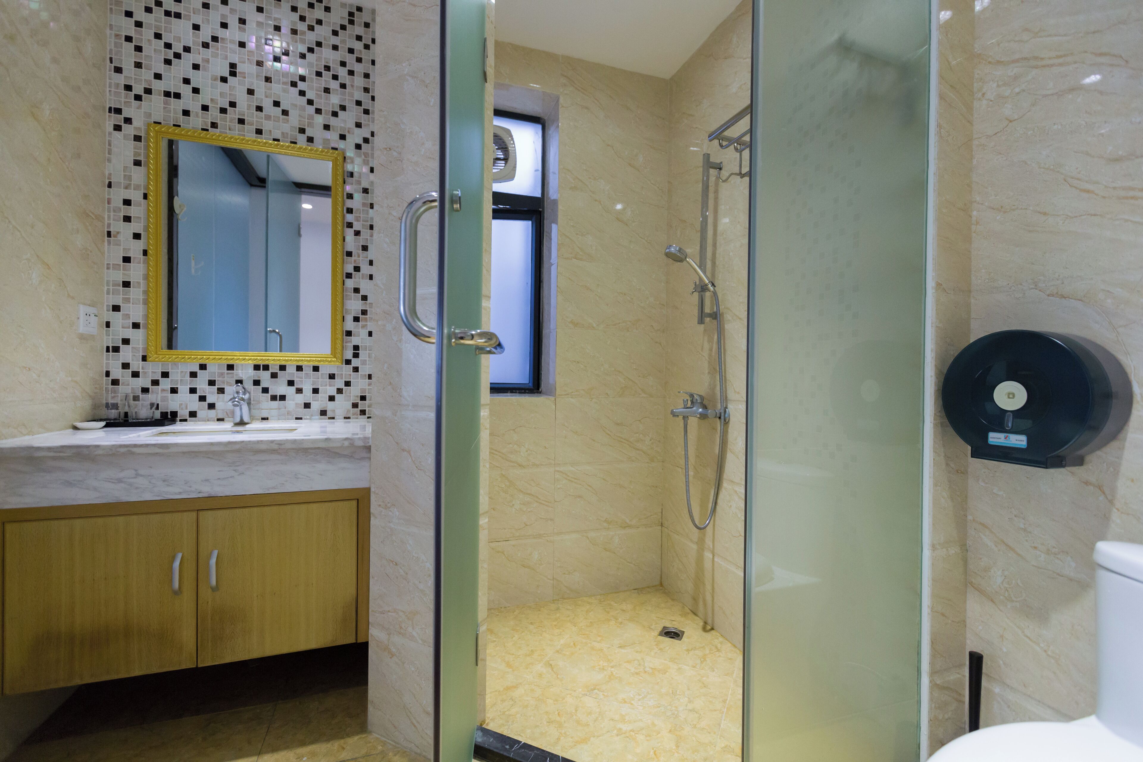 A bathroom with a glass-enclosed shower, a marble-tiled wall, a sink with a gold-framed mirror, and a toilet with a black toilet paper holder.