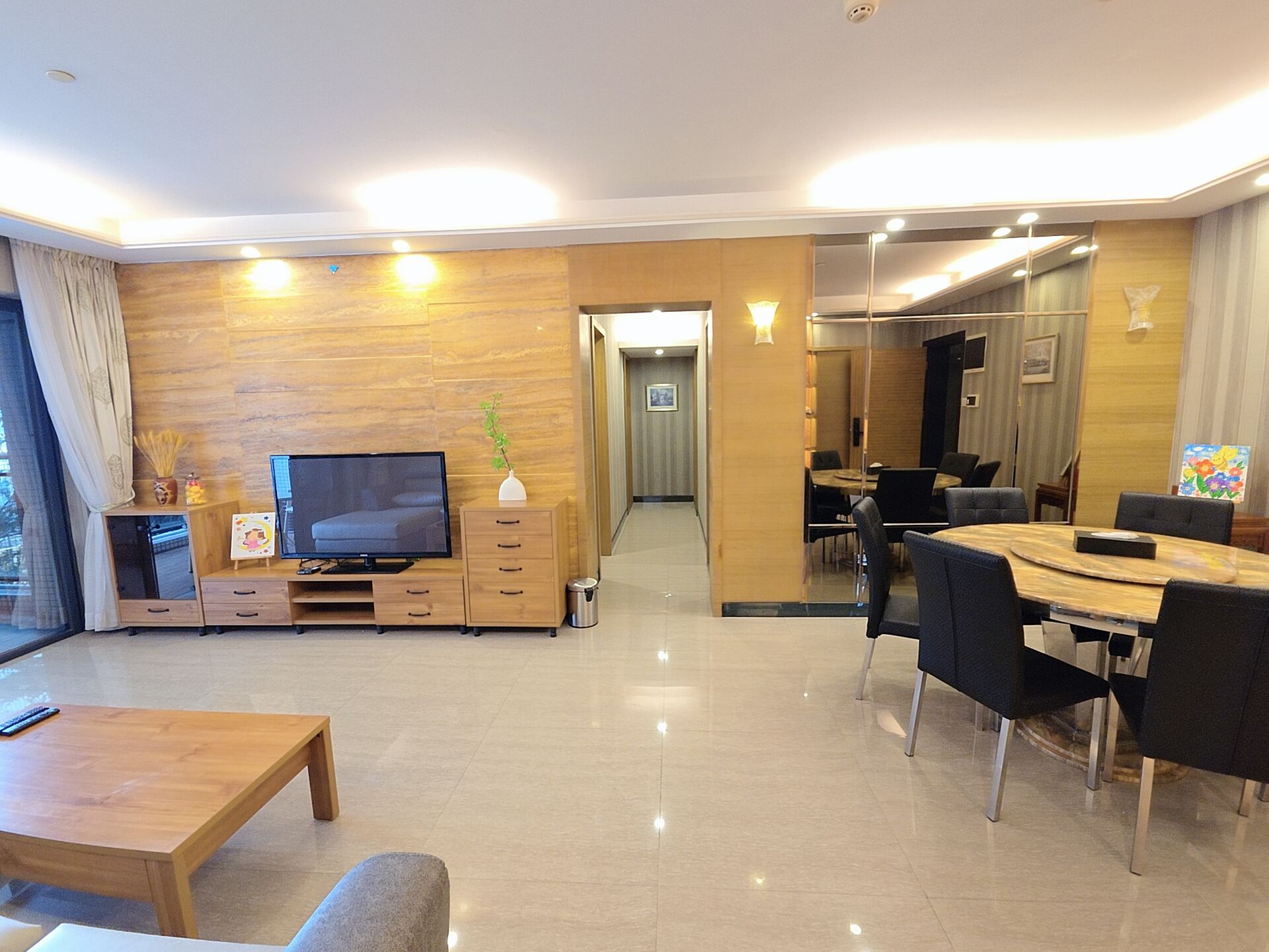 Una sala moderna con televisor de pantalla plana, muebles de madera, una zona de comedor con una mesa redonda y sillas, y un pasillo que conduce a otra habitación.
