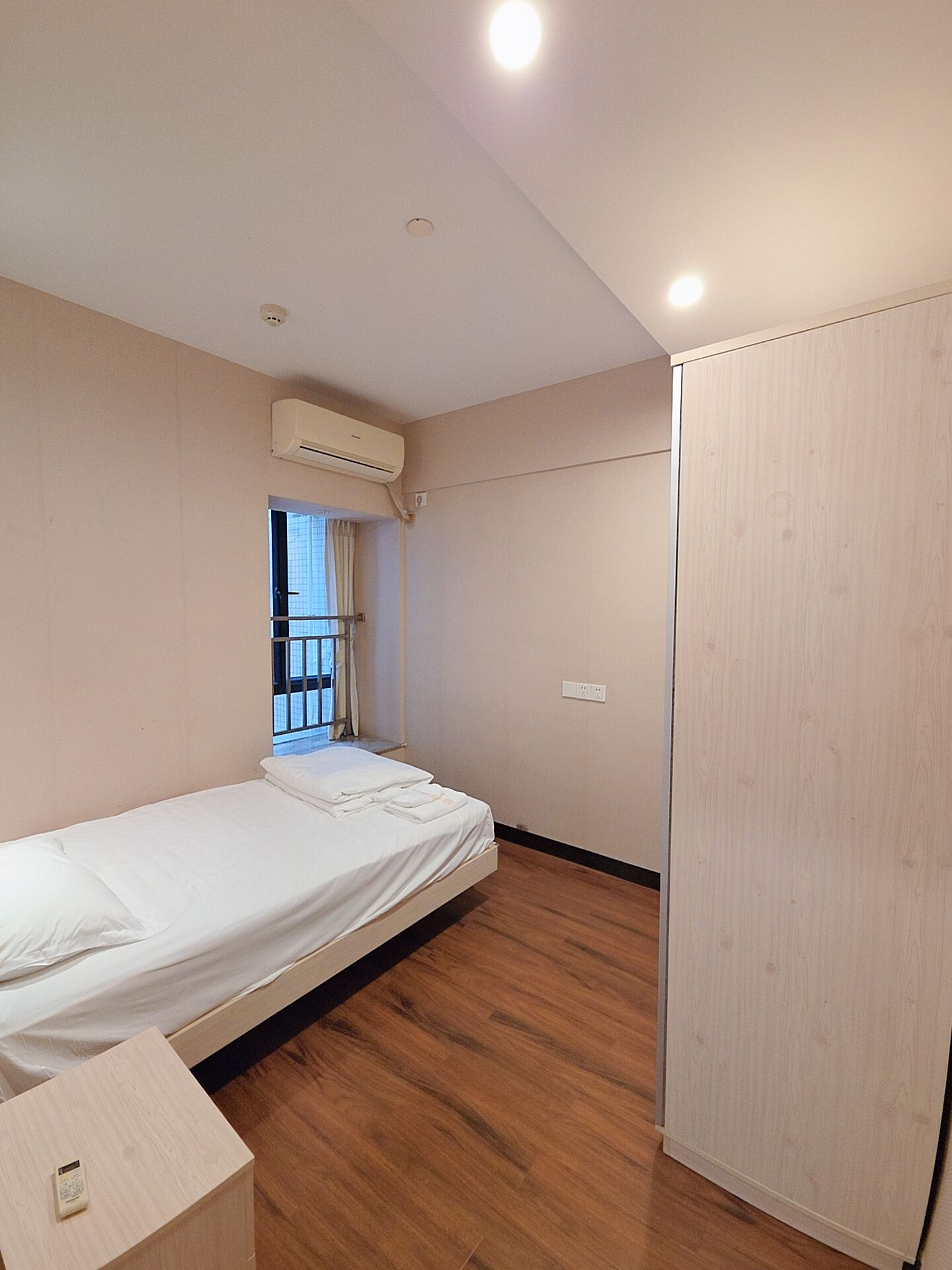 A single-bed room with a wooden floor, a window with curtains, an air conditioning unit, and a wardrobe.