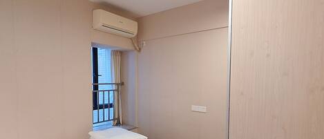 A single-bed room with a wooden floor, a window with curtains, an air conditioning unit, and a wardrobe.