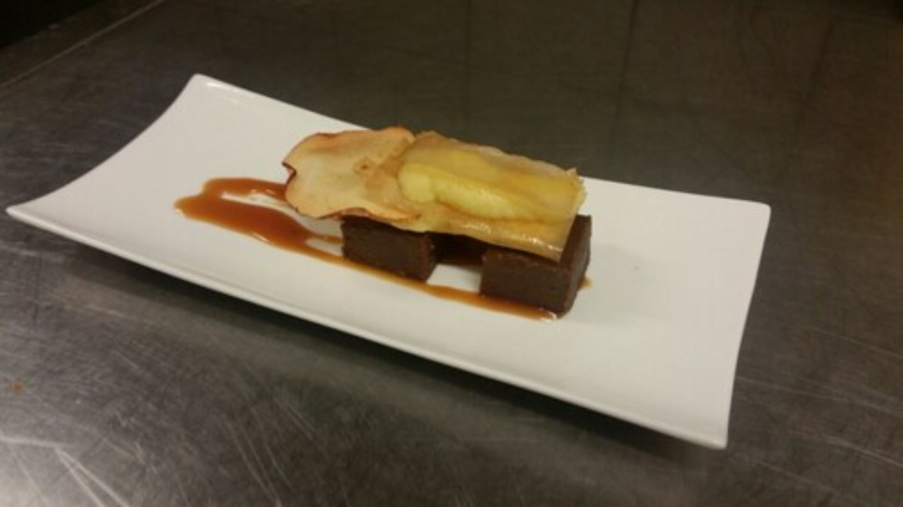 A rectangular white plate with a dessert consisting of a dark chocolate base, topped with a golden-brown pastry and a slice of what appears to be a fruit or pastry.