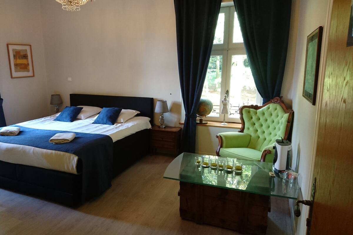 A neatly made bed with blue and white linens, a green armchair, a glass-top table with glasses, and a globe on a nightstand.