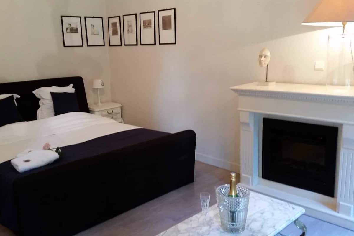 A bedroom with a black bed, white bedding, a bedside table, a lamp, a fireplace, and a framed photo collection on the wall.