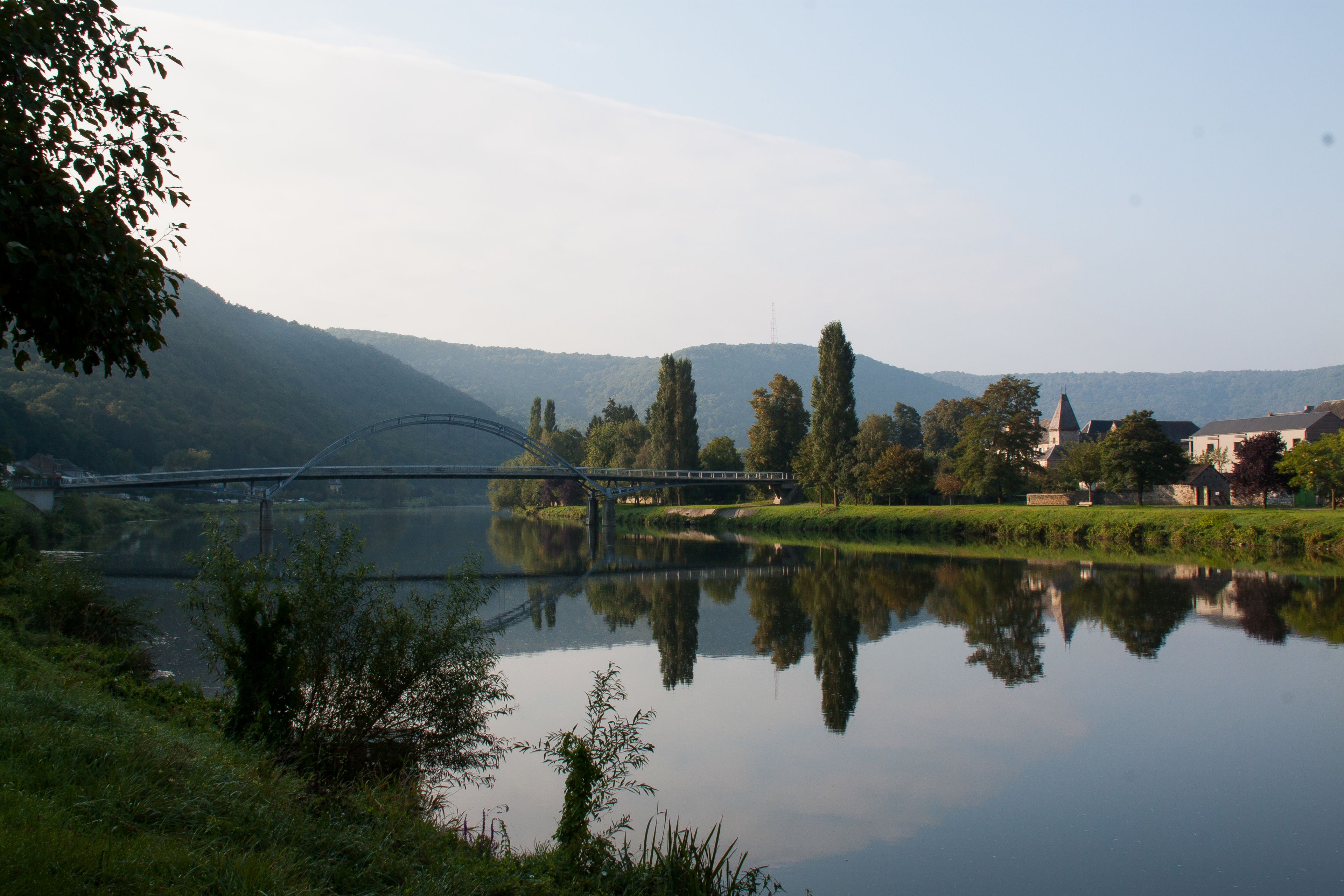 Un riu tranquil amb un pont, arbres i edificis al fons.