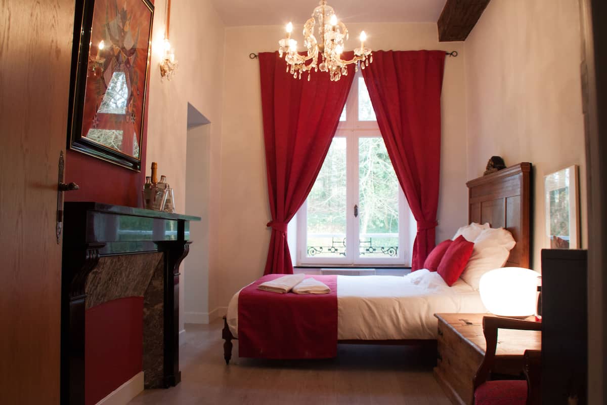 A bedroom with a bed, a wooden dresser, a chandelier, and a window with red curtains.