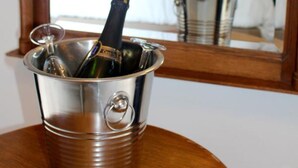 Un balde de metal con una botella y un vaso sobre una mesa de madera, reflejando un dormitorio con una cama, almohadas y una lámpara al fondo.