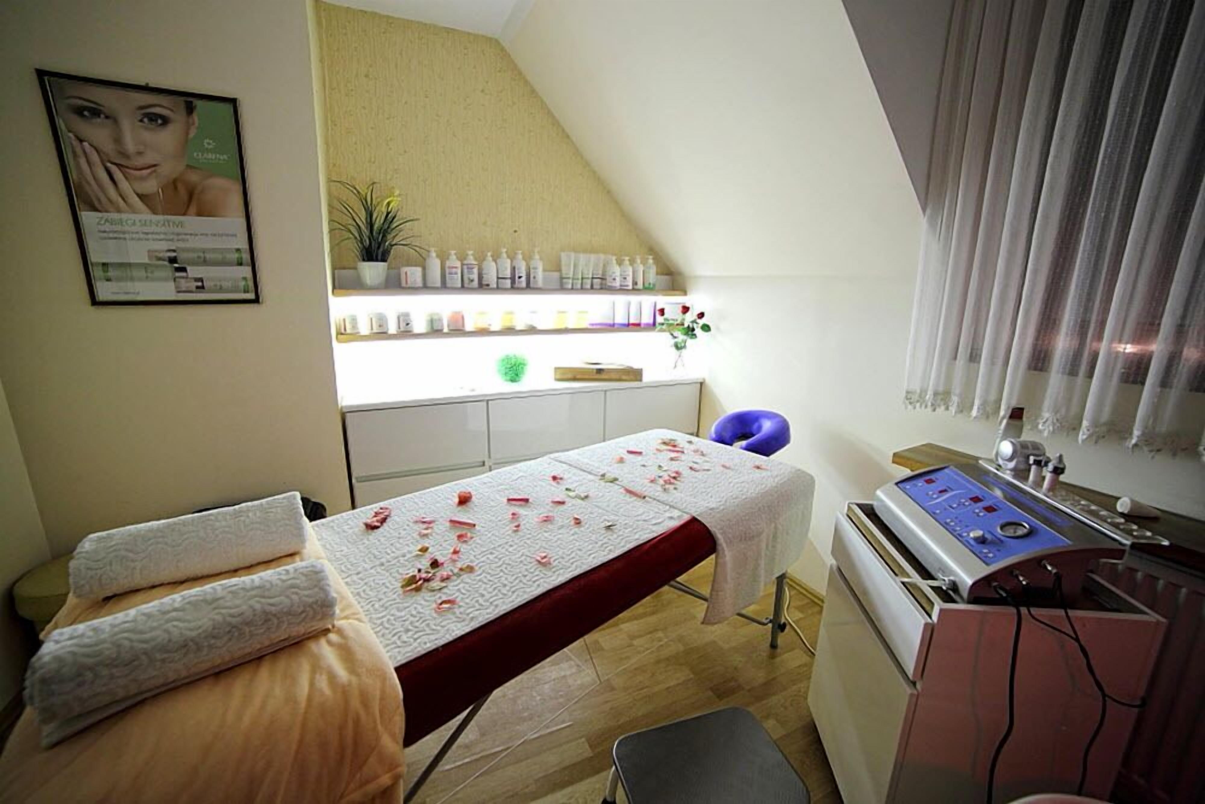A treatment room with a bed, a massage table, a shelf with bottles, a plant, and a massage machine.