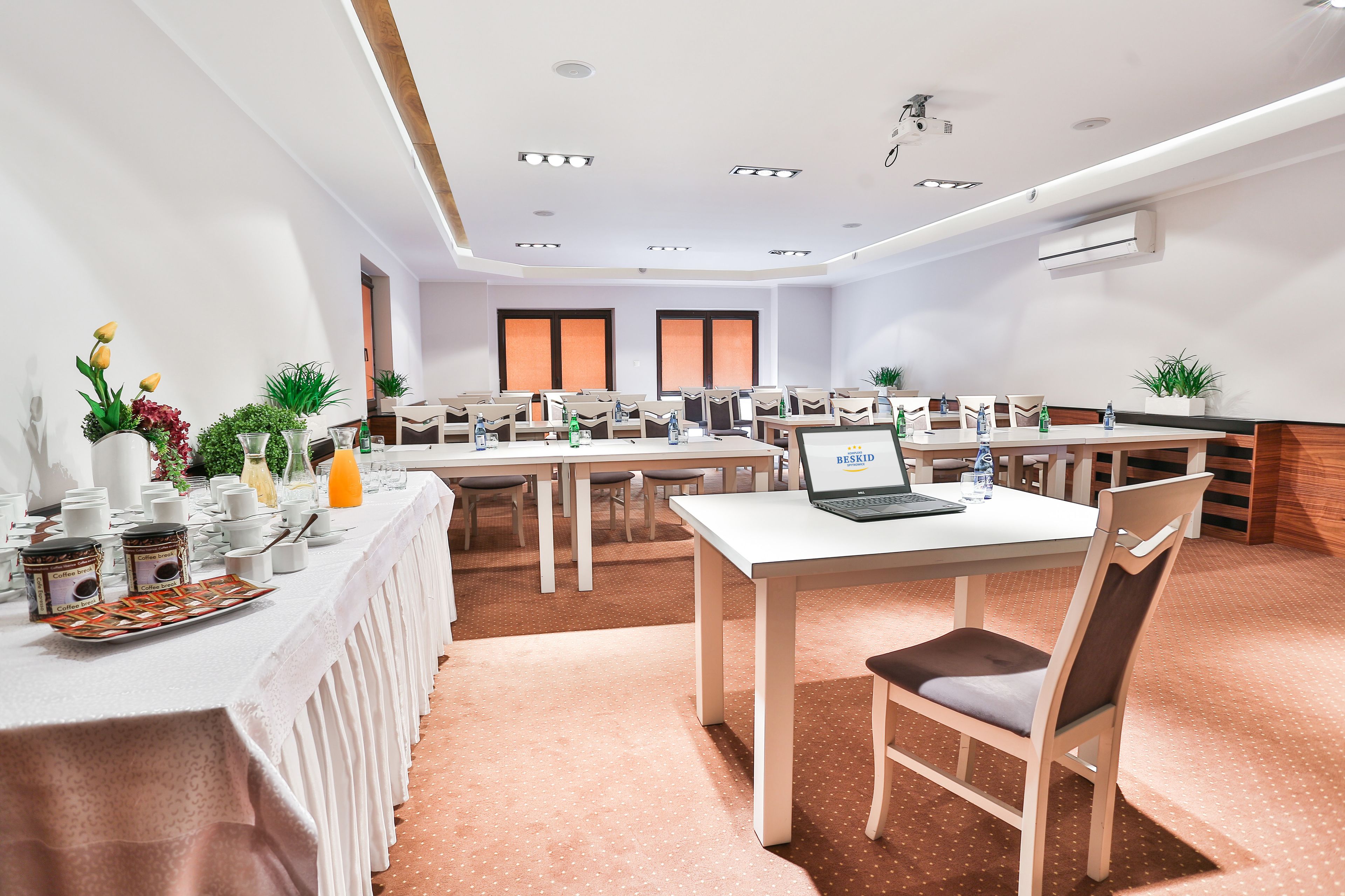 A conference room with white tables, chairs, and a laptop. There are potted plants and a tray of food on the tables.