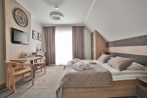 Quarto de hotel moderno com cama, escrivaninha, poltrona, televisão e uma janela grande com cortinas.