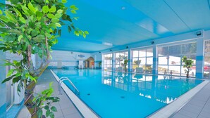 An indoor swimming pool with clear blue water, surrounded by large windows offering views of the outside.