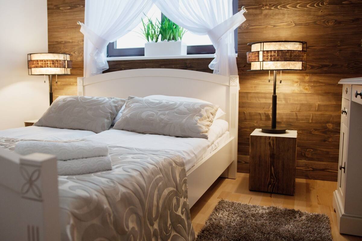 Un dormitorio ordenado con cama, lámparas de noche, una mesita de noche de madera y una ventana con una planta.