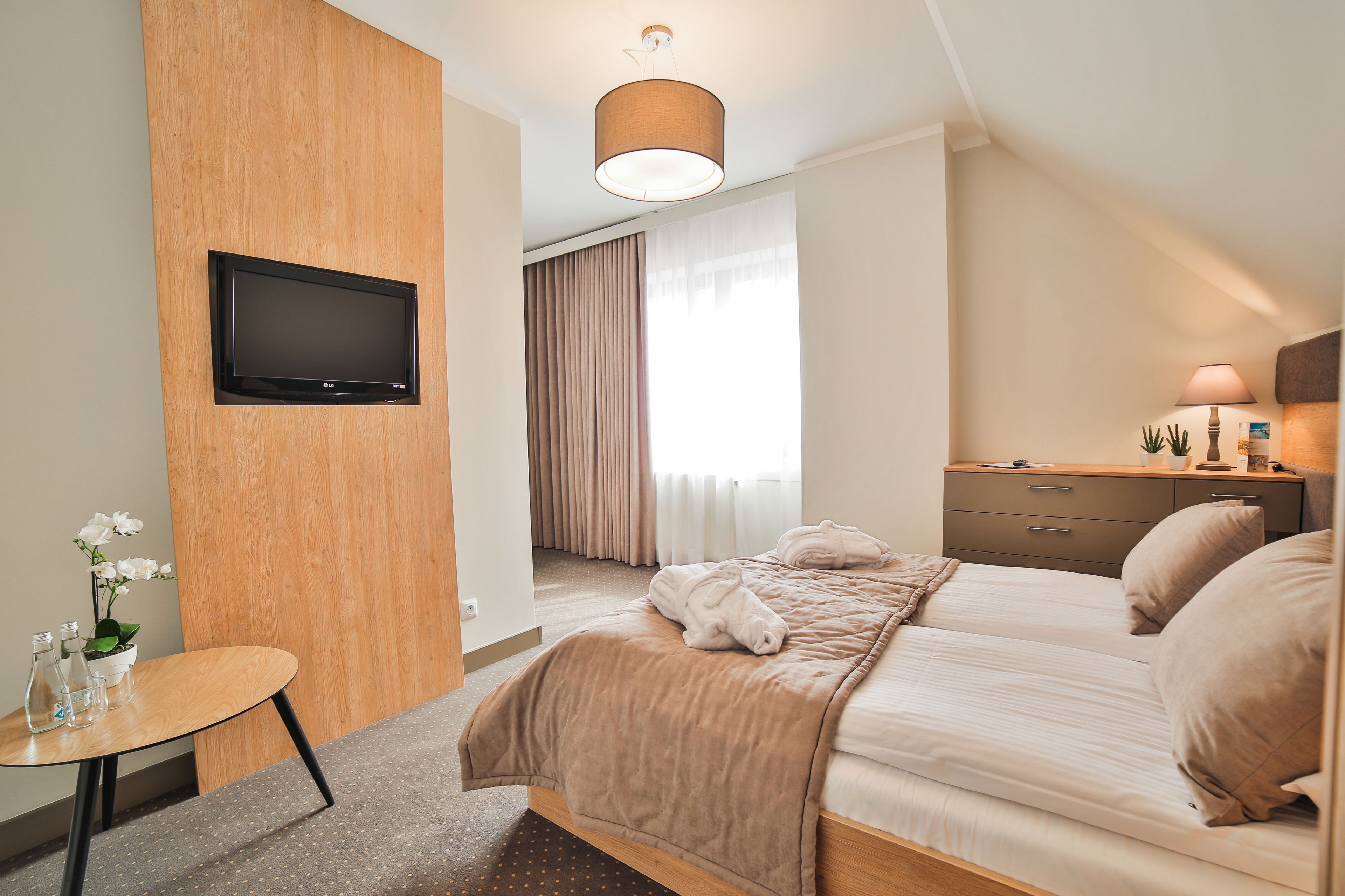 A hotel room with a bed, a TV mounted on the wall, a bedside table with a lamp, and a window with curtains.