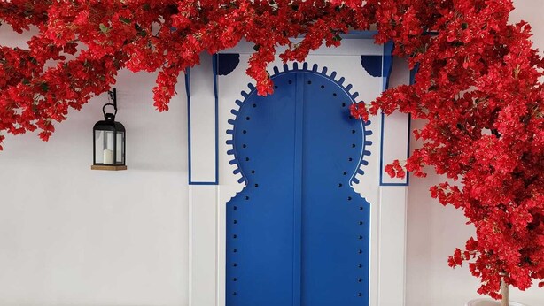 Eine blaue Tür mit einem dekorativen Metalldekor, eine weiße Bank und ein Blumentopf mit roten Blüten.