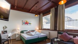 A hotel room with a bed, a table with a bottle and glasses, a chair, a painting, and a view of mountains through the window. - Gasthof Waldcafe (Mayrhofen)