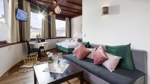 A cozy hotel room with a wooden ceiling, a sofa with green and pink pillows, a small table with a vase and glass, and a TV on the left wall. - Gasthof Waldcafe (Mayrhofen)