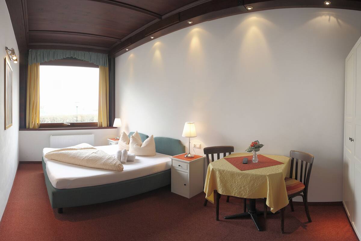 A hotel room with a bed, bedside table, lamp, chair, and a small table with a tablecloth.