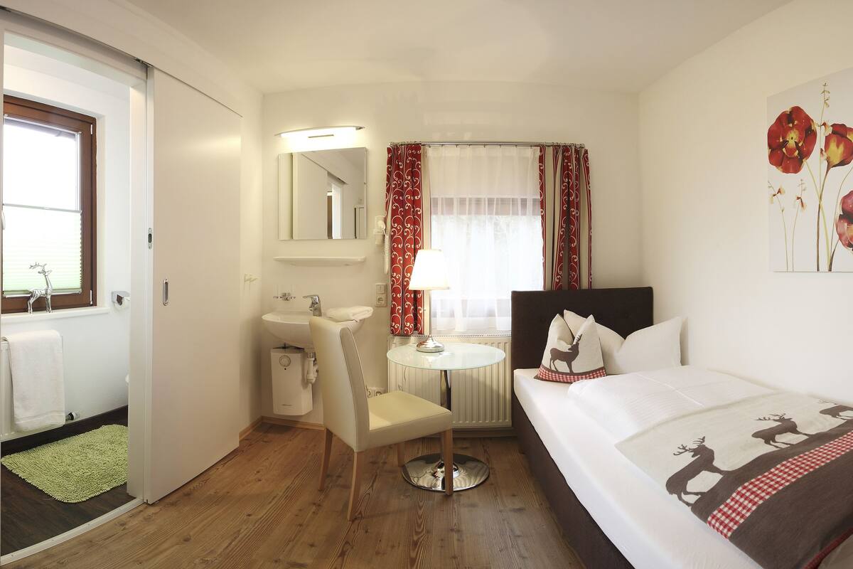 A hotel room with a bed, a round table with a lamp, a chair, a bathroom with a sink and mirror, and a window with curtains.