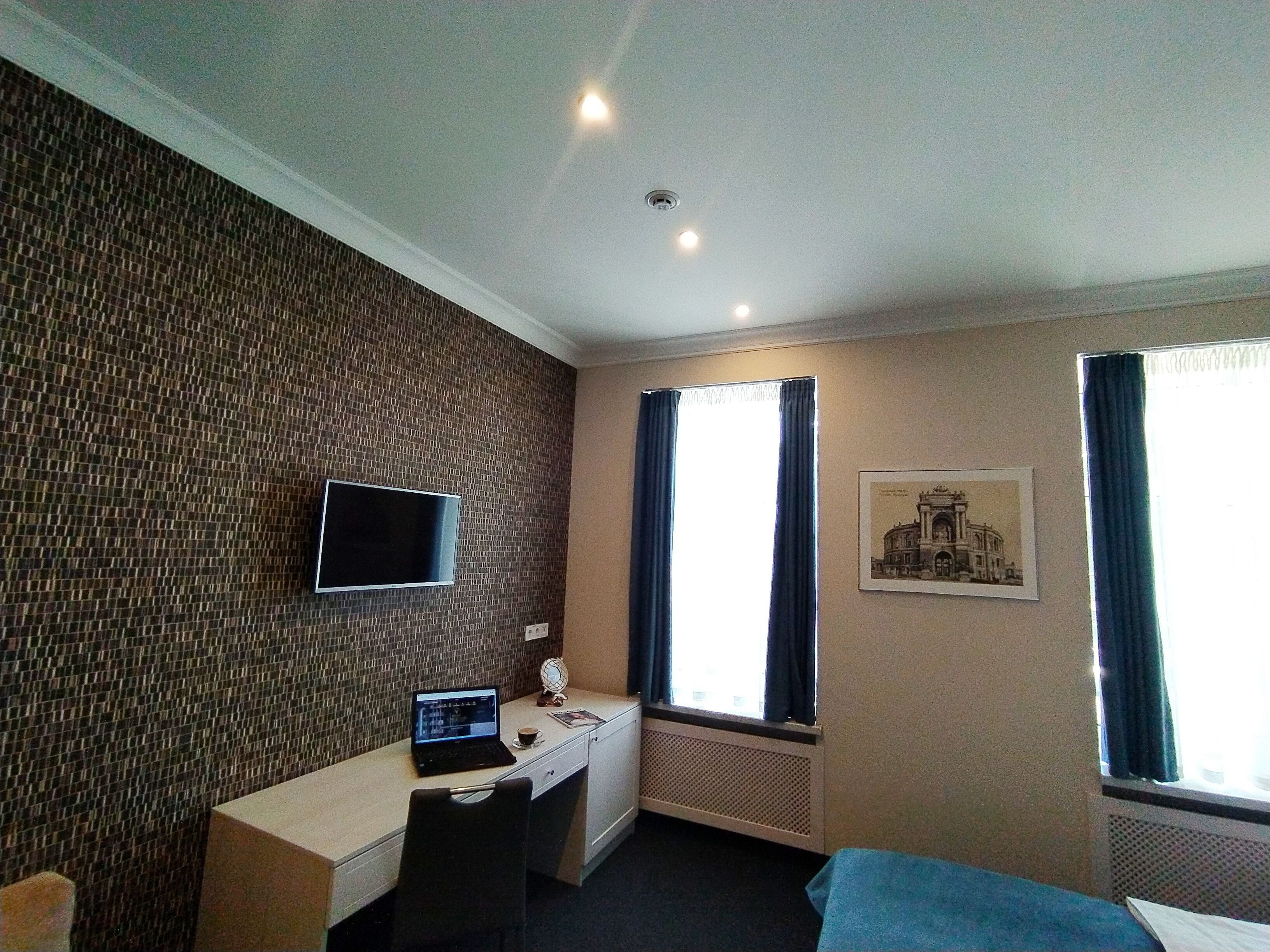 Habitación de hotel con pared de azulejos, televisor de pantalla plana, escritorio con computadora portátil, silla, cama y dos ventanas con cortinas.