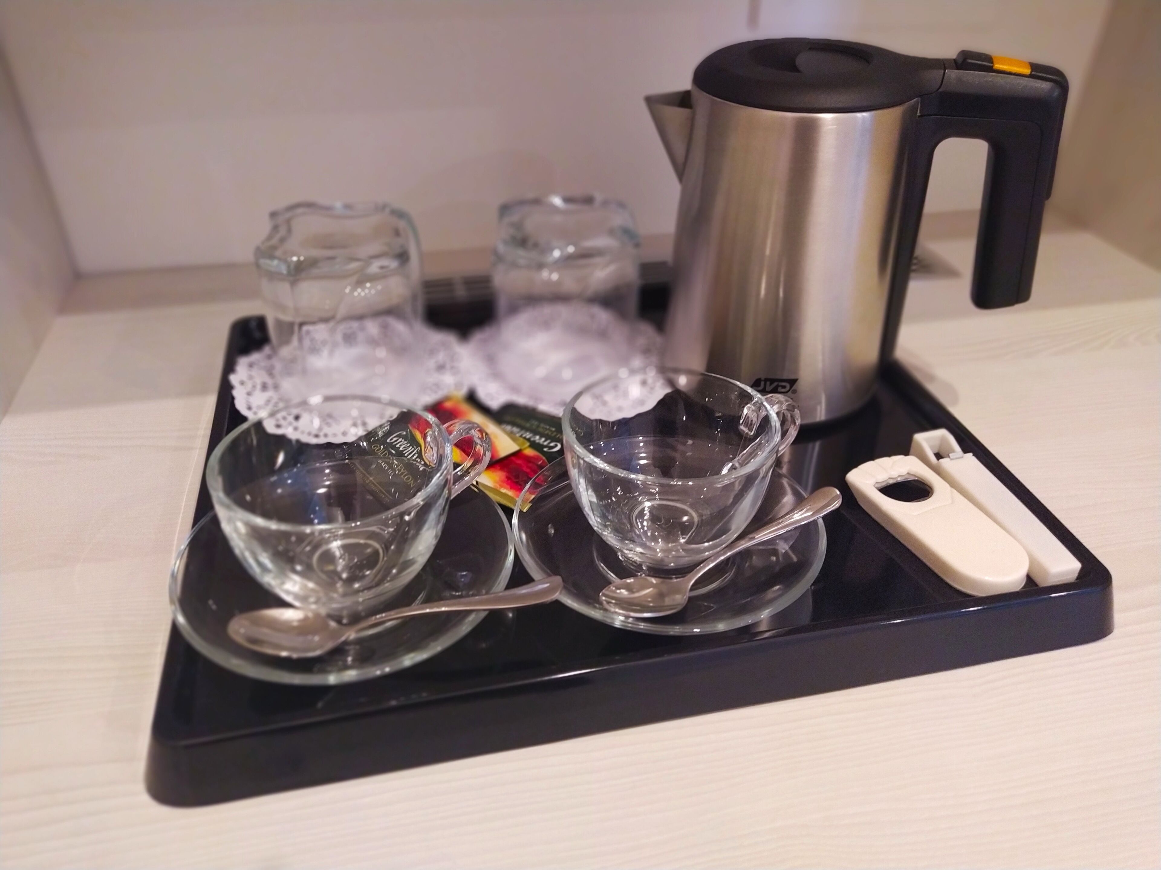 A tray with four clear glass cups, a silver teapot, and a small white device on a black tray.