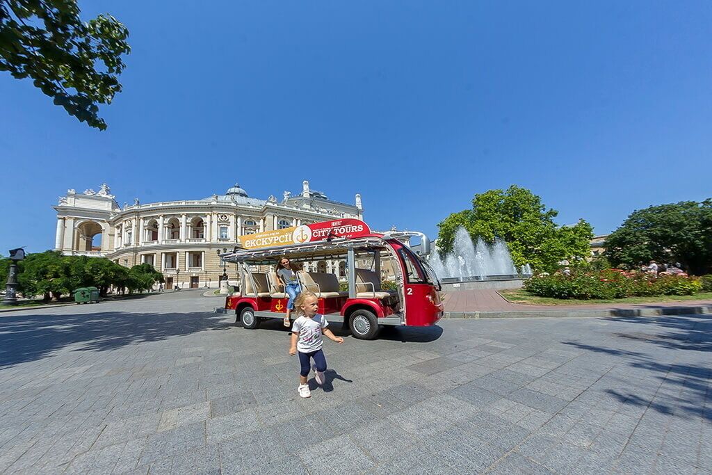 Un niño caminando frente a un autobús turístico rojo, con un edificio al fondo.