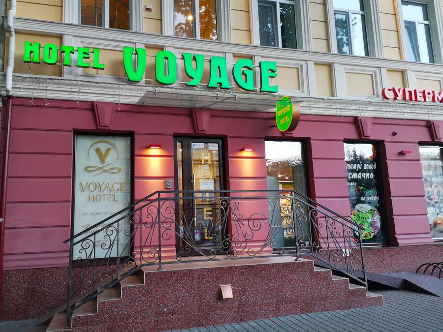 Un hotel llamado "HOTEL VOYAGE" con un letrero llamativo, una escalera que conduce a la entrada y una puerta visible con un letrero que indica "CAMPING" y "Fruta fresca".
