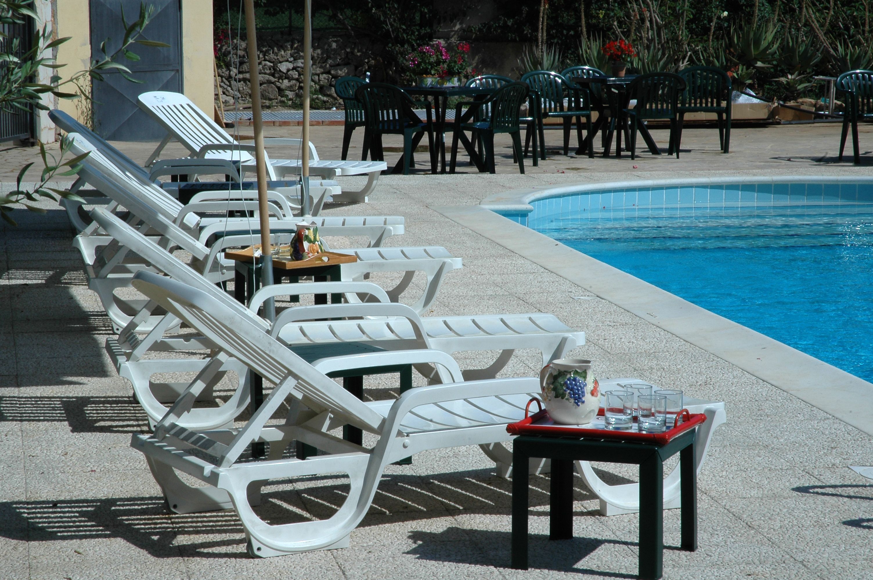 Un espace aménagé au bord de la piscine, avec des chaises longues et des tables blanches, une petite table sur laquelle sont posés une carafe et des verres, et une piscine.