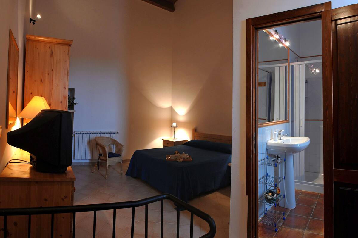 Ein Schlafzimmer mit Bett, Nachttisch, Lampe, Stuhl und Blick auf ein Badezimmer mit Waschbecken und Spiegel.