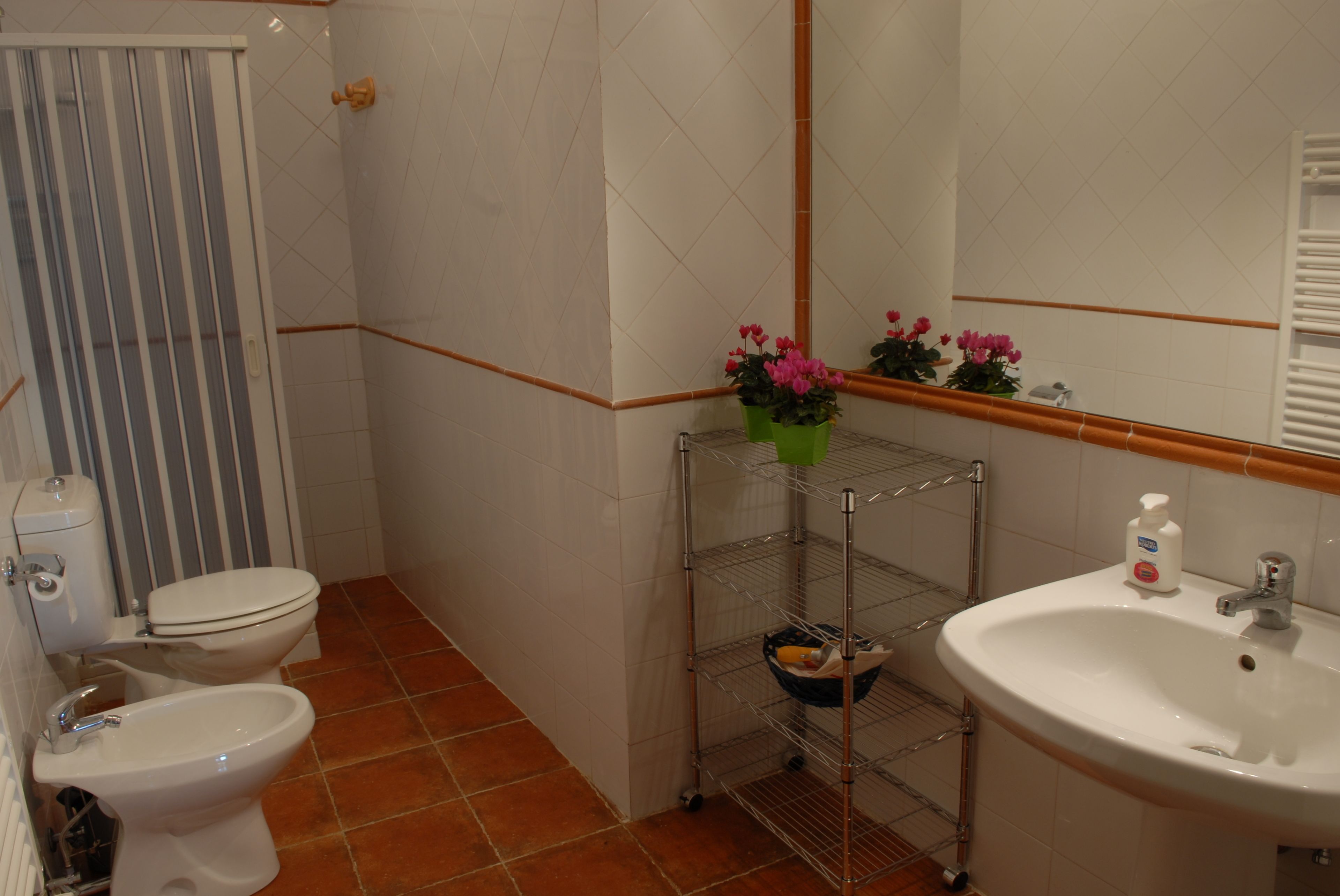 A bathroom with a toilet, bidet, sink, and a shelf with flowers.