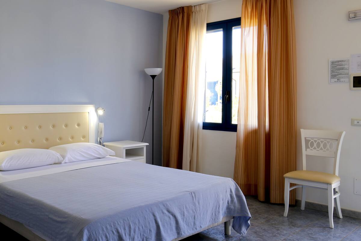 A bedroom with a bed, a chair, a nightstand, a lamp, and a window with curtains.