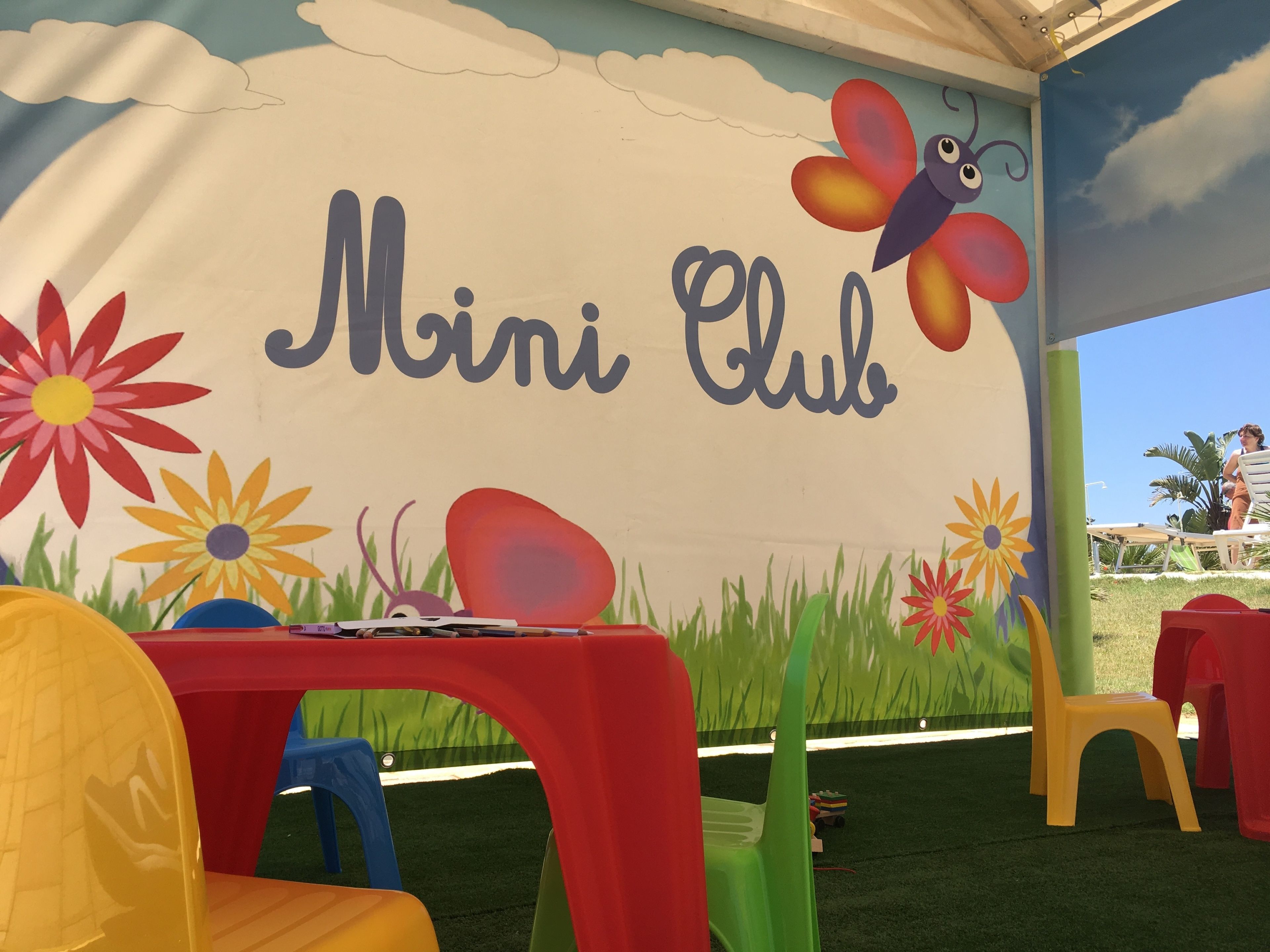 Un'area giochi per bambini con un cartello 'Mini Club', sedie colorate e un tavolo pieno di giochi.