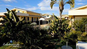 A resort with multiple yellow buildings, lush tropical plants, and a clear blue sky. - Calabernardo Resort (Noto)