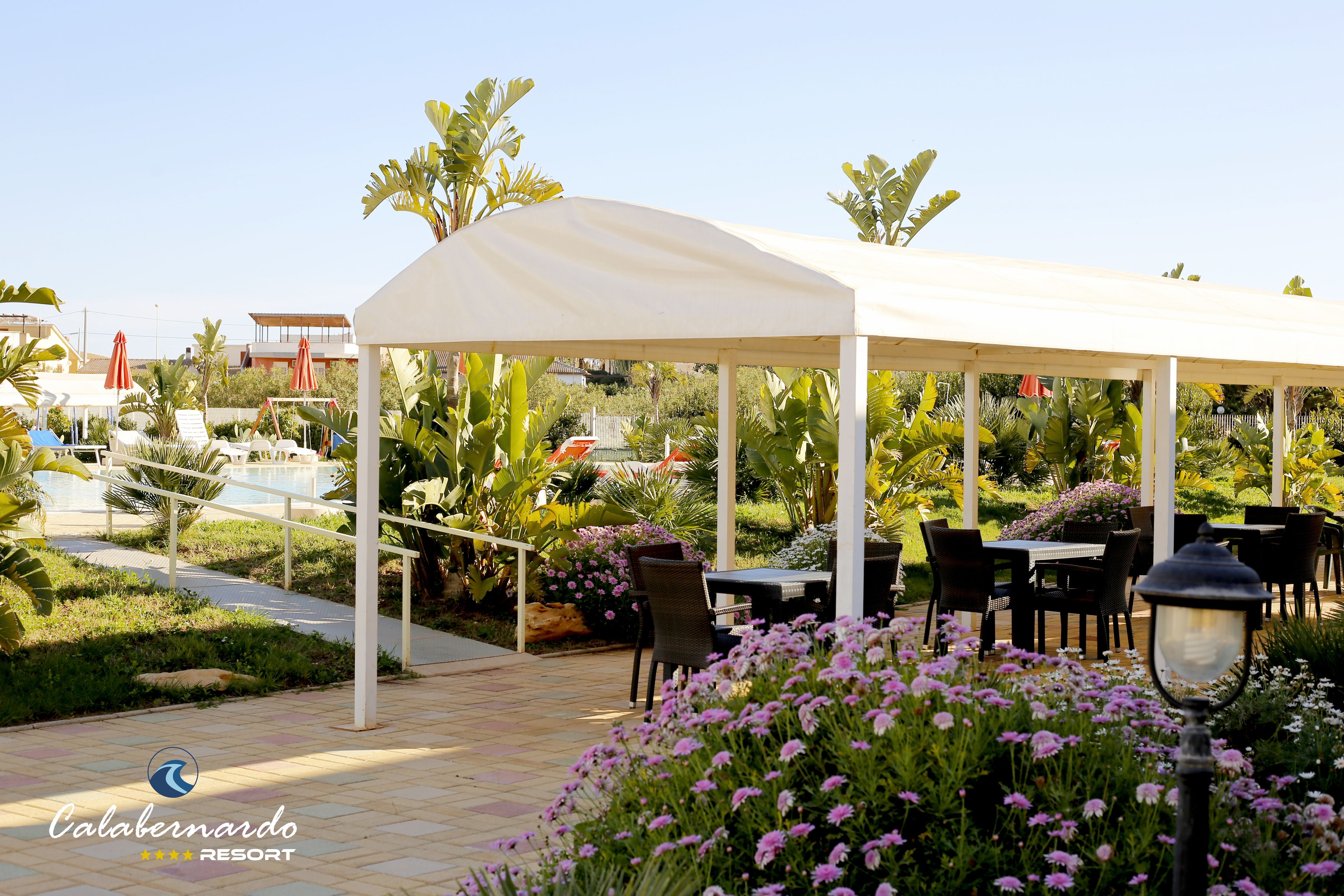 A covered outdoor dining area with tables and chairs, surrounded by lush greenery and flowers.