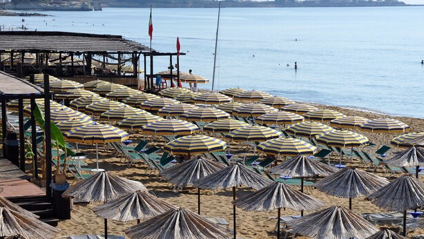 A beach with rows of sun umbrellas and lounge chairs, a covered area with tables and chairs, and a view of the sea and distant hills. - Calabernardo Resort (Noto)