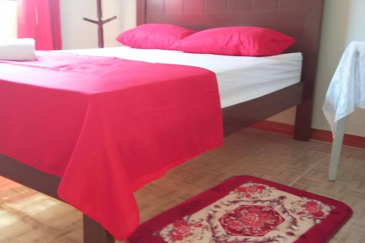 A bed with a red bedspread and white linens, a wooden headboard, and a red and white patterned rug on the floor.