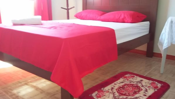 A bed with a red bedspread and white linens, a wooden headboard, and a red and white patterned rug on the floor.