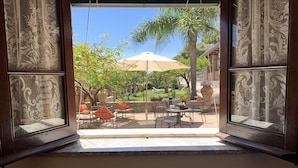 Uitzicht door een raam met gordijnen met een patroon, dat een terras buiten toont met tafels, stoelen en een grote parasol.