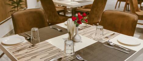 A dining area with tables set for four, each with a placemat, cutlery, and a vase with red flowers.