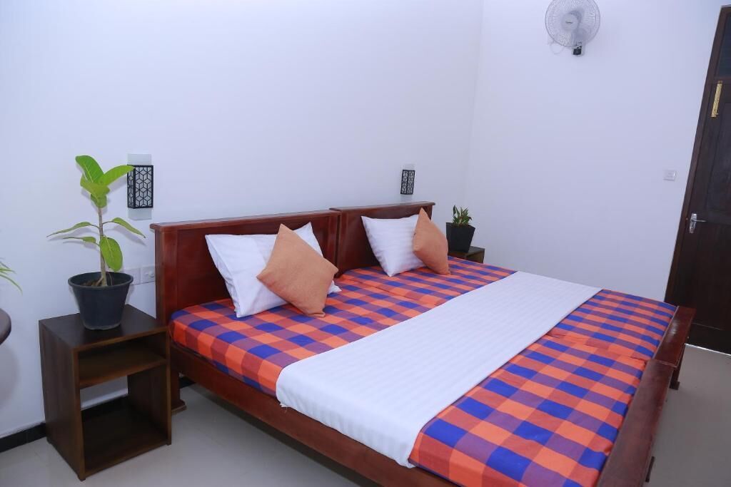 A single bed with a checkered bedspread, two white pillows, and a single orange pillow. There is a small wooden nightstand with a potted plant beside the bed. A wall-mounted fan is visible on the wall above the bed.
