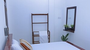 Una cama bien hecha con sábanas blancas y almohadas a rayas, cabecera de madera, ventilador fijado a la pared, una pequeña estantería con plantas y un espejo.