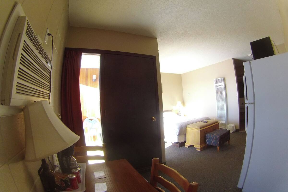 Habitación de hotel pequeña con kitchenette, una cama, una mesita con lámpara y puerta a un balcón.