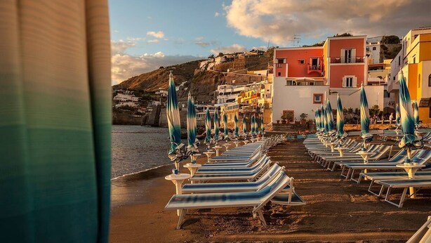 A beach with rows of sun loungers and umbrellas, a hillside with colorful buildings, and a clear sky.